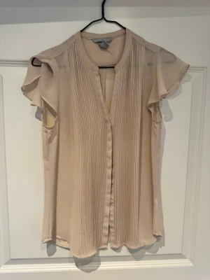 Beige plisserad blus från H&M - Snygg beige blus från H&M med plisserade detaljer framtill och korta, volangiga ärmar. Blusen har en lätt och luftig känsla, knäppning framtill och är tillverkad i ett tunt, mjukt tyg som ger ett elegant intryck.