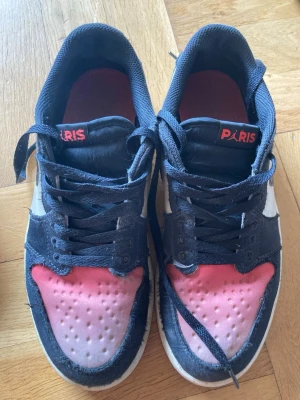 Nike Paris Saint Gemain  svarta och rosa sneakers str. 40,5 - Snygga Nike sneakers med svart ovandel, vita detaljer och rosa tåparti. På plösen står det 'PARIS' i rött. Skorna har klassisk snörning och röd innersula. Perfekta för dig som gillar streetstyle och unika färgkombinationer. Köpta i Paris Saint Gemain butiken i New York. Storlek 40,5 (25,5 cm). Original skokartong medföljer. 
