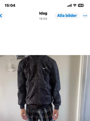 Svart Nike vindjacka med camomönster - Snygg svart vindjacka från Nike med diskret camomönster och vit logga på bröstet. Jackan har dragkedja framtill, resår vid ärmslut och midja samt två sidofickor med dragkedja. Perfekt för en sportig och avslappnad stil.