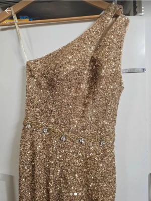 Glittrig guldig one shoulder klänning - En glittrig långklänning i guld med paljetter och dekorativt bälte i midjan. Klänningen har en axelbandslös design med one shoulder och är perfekt för speciella tillfällen. Materialet är troligtvis polyester eller syntet.