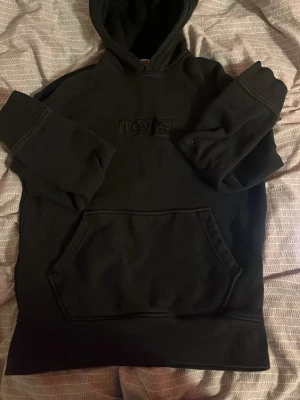 Svart hoodie från Levi's med broderad logga - Svart hoodie från Levi's med broderad logga på bröstet. Klassisk känguruficka framtill och huva. Mjuk bomullskänsla och ribbade muddar vid ärmslut och nederkant. Perfekt för en avslappnad och stilren look.