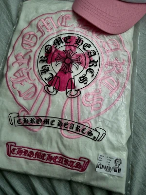 Chrome Hearts långärmad topp rosa/vit - Säljer en långärmad topp från Chrome Hearts i vitt med ljusrosa ärmar och tryck. Toppen har ett stort Chrome Hearts-tryck i rosa och svart på ryggen och ett mindre tryck på bröstet. Skön och avslappnad passform, perfekt för en cool streetwear-look.