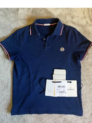 Marinblå Moncler pikétröja - Snygg marinblå pikétröja från Moncler med klassisk krage och knappar framtill. Tröjan har vita och röda detaljer på krage och ärmslut samt Moncler-logga på bröstet. Perfekt för en stilren och trendig look.