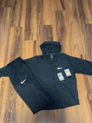 Nike miller tracksuit - Snyggt svart träningsset från Nike med matchande jacka och byxor. Jackan har huva och dragkedja framtill, båda plaggen har den klassiska vita Nike-loggan. Perfekt för träning eller chill. Materialet är lätt och andas.