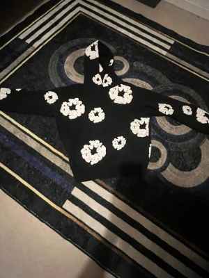Svart hoodie med vita blommor - Cool svart hoodie med stora vita blommor tryckta över hela tröjan, även på huvan och ärmarna. Perfekt för dig som vill sticka ut med en unik och trendig stil. Mjuk och skön, passar till många outfits.