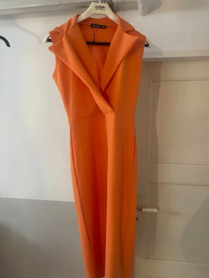 Orange långklänning från Boohoo - Snygg orange långklänning från Boohoo med omlottkrage och ärmlös design. Klänningen har en stilren och modern look, perfekt för dig som vill sticka ut. Tillverkad i ett mjukt syntetmaterial som ger en skön känsla.