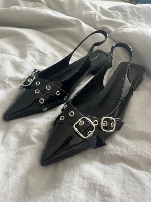 Svarta lackade ballerinaskor med nitar - Snygga svarta ballerinaskor i lack från din sko med spetsig tå, remmar med silverfärgade nitar och spännen. Skorna har öppen häl och en låg klack, vilket ger en edgy och trendig look. Perfekta för dig som vill sticka ut lite extra. Aldrig använt 