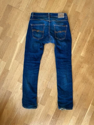 Mörkblå Grim Tim Nudie Jeans 32/34 - Säljer ett par mörkblå grim tim jeans från Nudie Jeans. De har riktigt skön stretch, klassiska fem fickor och snygga slitningar på framsidan. Jeansen har lagats av Nudie i skrevet. Jeansen har den klassisk Nudie-loggan på bakfickan. Perfekta för en avslappnad och trendig look. Storlek 32/34