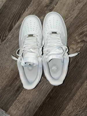 Nike Air Force 1 vita sneakers - Klassiska Nike Air Force 1 sneakers i helvitt läder med vit sula och vita skosnören. Ikonisk design med perforerad tåbox och Nike Swoosh på sidan. Perfekta för dig som gillar stilrena och tidlösa sneakers.