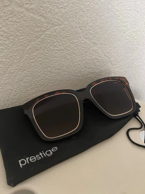 Bruna Prestige solglasögon med gulddetalj - Snygga solglasögon från Prestige med fyrkantig form och brunt sköldpaddsmönster på bågarna. Glasen är mörka och bågarna har en tunn guldfärgad detalj runt glasen. Perfekta för att lyfta din stil i solen.