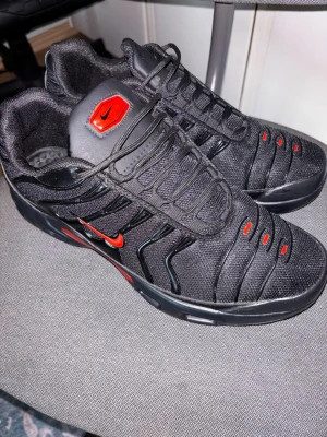 Nike svarta sneakers med röda detaljer - Svarta Nike sneakers med röda detaljer och logga. Skorna har en sportig design med mesh och syntetmaterial, samt svarta snören och röda accenter på tungan och sidorna. Perfekta för dig som gillar en stilren och modern look.