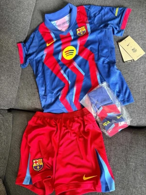 FC Barcelona matchställ Nike  - Säljer ett FC Barcelona matchställ från Nike i storlek XL för barn. Tröjan är blå med röda och gula detaljer, klubbmärke och sponsorlogga. Shortsen är röda med klubbmärke och Nike-logga. Setet inkluderar även matchande strumpor. Perfekt för fotbollsträning eller fans.