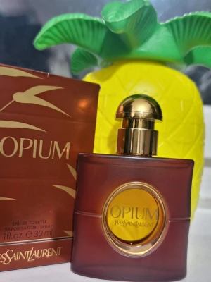 Opium parfym från Yves Saint Laurent - Opium Eau de Toilette från Yves Saint Laurent i en elegant brunröd glasflaska med gulddetaljer och rund gul etikett. Flaskan rymmer 30 ml och har en lyxig, klassisk design som känns både tidlös och exklusiv.