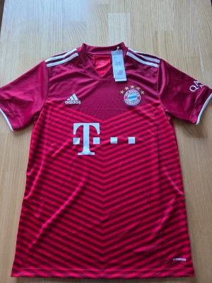 Adidas FC Bayern München 21/22 fotbollströja str. M - Snygg röd Bayern München matchtröja från Adidas med vita detaljer och klubbmärke på bröstet. Tröjan har AEROREADY-material som andas, korta ärmar och klassiska ränder på axlarna. Perfekt för fotbollsträning eller att bära på läktaren.