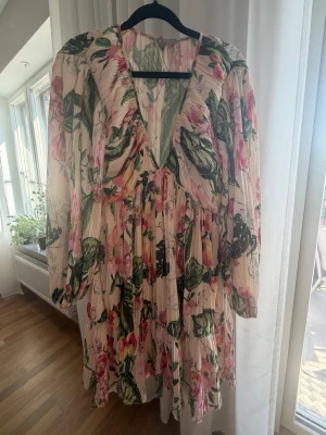 Blommig v-ringad klänning med volang - Romantisk blommig klänning i ljusa toner med gröna och rosa detaljer. Klänningen har v-ringning, långa ärmar och volangdetaljer. Plisserad design ger ett luftigt intryck och tyget känns lätt och flowy, perfekt för varmare dagar.