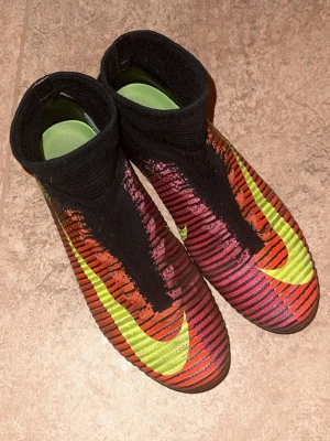 Nike Mercurial superfly 5  - Säljer ett par Nike Mercurial superfly 5 fotbollsskor med hög strumpa. Skorna har en färgstark design i rött, lila/rosa och svart med stora neongula swoosh-loggor. Ovandelen är ribbad för bättre bollkontroll och sulan har fasta dobbar för spel på gräs. Pris kan diskuteras 
