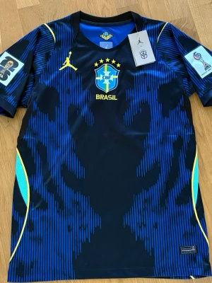 Brasilien away 2026 - Snygg blå och svart matchtröja från Brasilien med gula detaljer och CBF-logga på bröstet. Tröjan har korta ärmar, rund hals och är tillverkad i ett lätt, ventilerande funktionsmaterial. Perfekt för fotboll eller träning.