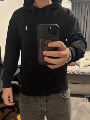 Svart hoodie från Polo Ralph Lauren - Svart hoodie från Polo Ralph Lauren med klassisk huva och dragsnören. Liten vit broderad logga på bröstet och känguruficka framtill. Mjuk bomullskänsla och ribbade muddar vid ärmslut och midja. Perfekt för en avslappnad och stilren look.                         Längd: 65 cm.                                                   Bröstvidd: 53 cm