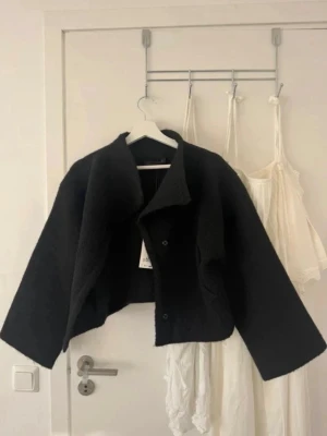 Svart oversized cardiganjacka Zara - Snygg svart cardiganjacka från Zara med oversized passform och breda ärmar. Jackan har en kort, boxig siluett, stora slag och dolda knappar framtill. Tillverkad i ett mjukt, ull-liknande material som ger en cool och modern look.