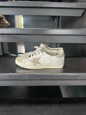 Golden goose sneakers  - Snygga vita sneakers från Golden Goose med en glittrig stjärna på sidan. Skorna har lågt skaft, vita snören och detaljer i mocka och textil. Perfekta för dig som vill ha en cool och trendig look med lite extra bling. Sulan är omgjord och helt ny då den var sönder i hälarna 