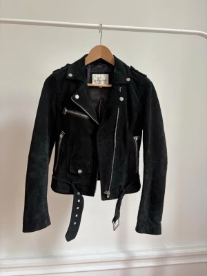 mockajacka från review - Snygg svart mockarjacka i bikerstil från review med silvriga dragkedjor, axelklaffar och bälte i midjan. Jackan har flera fickor med dragkedja och klassisk krage. Perfekt för en cool och edgy look.