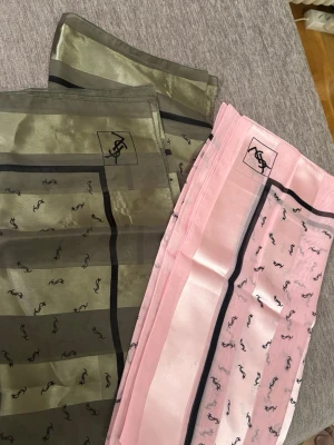 YSL sidenscarf i rosa och olivgrönt - Två lyxiga sidenscarves från Yves Saint Laurent, en i olivgrönt och en i ljusrosa. Båda har svarta ränder och YSL-loggan tryckt i hörnet samt små loggor över hela tyget. Perfekt accessoar för att lyfta din outfit.