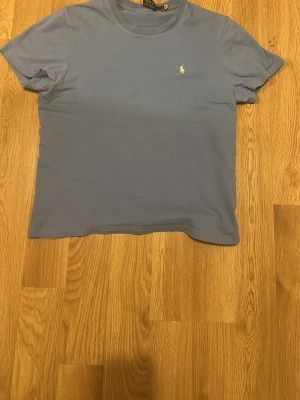 Blå t-shirt från Ralph Lauren - Snygg blå t-shirt från Ralph Lauren med klassisk passform och liten broderad logga på bröstet. T-shirten är kortärmad och tillverkad i mjuk bomull, perfekt för en chill och stilren look. Passar de som är runt 182