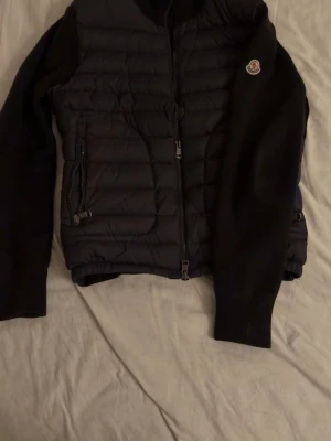 Svart quiltad jacka från Moncler - Snygg svart quiltad jacka från Moncler med dragkedja framtill och två sidofickor med dragkedja. Jackan har långärmade stickade ärmar och klassisk Moncler-logga på armen. Perfekt för dig som vill ha en stilren och bekväm jacka. Finns NFS märke i loggan. OBS! Oklart om riktig eller inte!