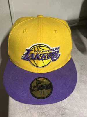 Lakers keps 7 3/8. 58,7cm - Snygg gul och lila keps från New Era med Los Angeles Lakers broderad logga framtill och NBA-märke bak. Flat skärm och klassisk 59FIFTY-modell. Perfekt för dig som vill visa ditt Lakers-stöd med stil.