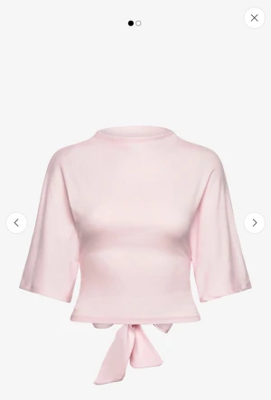 Ljusrosa topp med knyt och rosett - Supersöt ljusrosa topp med trekvartsärm och knyt i midjan som bildar en rosett i ryggen. Toppen har en croppad passform och är tillverkad i mjukt bomullsmaterial. Perfekt för dig som gillar en feminin och trendig stil.