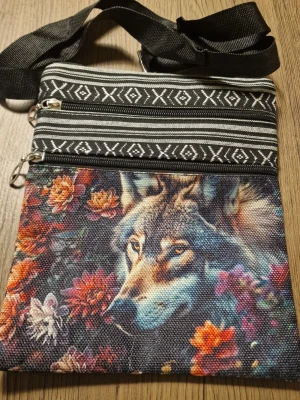 Axelväska med vargmotiv och blommor - Unik axelväska i tyg med färgglatt vargmotiv bland blommor på framsidan och svartvita etnomönster. Väskan har två dragkedjefack framtill och justerbar svart axelrem. Bredd 19 cm. Höjd 23,5 cm. Perfekt för dig som gillar djurmotiv och boho-stil.