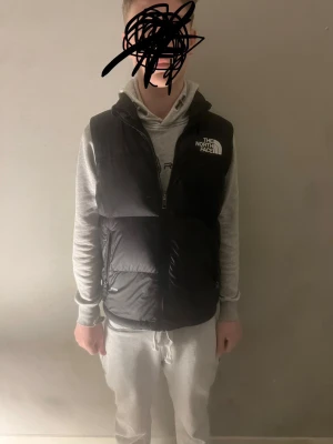 Svart dunväst från The North Face - Snygg svart dunväst från The North Face med vit logga på både bröstet och ryggen. Västen har dragkedja framtill och två fickor med dragkedja. Perfekt att slänga över en hoodie för en cool och sportig look. NY pris 2999kr så det är ett otroligt billigt pris så passa på! 
