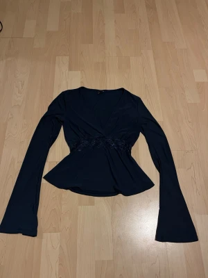 Mörkblå omlottopp med spetsdetalj - Snygg mörkblå omlottopp med v-ringning och långa ärmar. Toppen har en markerad midja med spetsdetalj som ger en feminin siluett. Perfekt att styla med jeans eller kjol för en trendig look.
