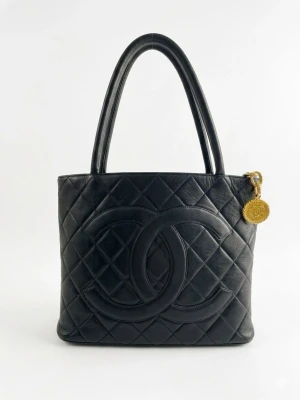 Chanel quilted Medallion Tote bag black  - Jag säljer min Chanel Medallion Tote i vacker svart lammnappa med den ikoniska stora CC-loggan framtill. Väskan är rengjord både på insidan och utsidan och lätt vaxad för skydd, men är obehandlad och ofärgad, vilket bevarar det ursprungliga skicket. Väskan är i mycket gott skick.  Väskan har en topplås med dragkedja och ett medaljongformat Chanel CC-dragkedjehandtag i guldfärgad metall. Den har sin originala serienummer (6352982) inuti, vilket motsvarar en produktionstid på cirka 2000–2002. 