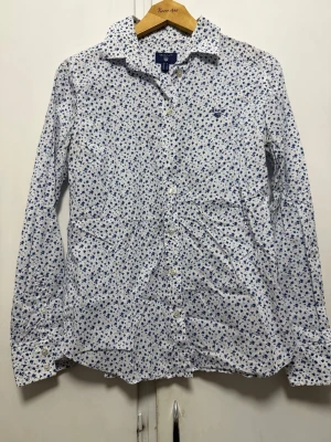 Vit blommig skjorta från Gant - Vit skjorta från Gant med små blå blommor över hela tyget. Klassisk krage, långa ärmar och knäppning framtill. Tillverkad i mjuk bomull som känns skön mot huden. Perfekt för dig som gillar en fräsch och stilren look med lite extra detaljer.