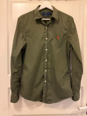 Olivgrön skjorta från Ralph Lauren - Klassisk olivgrön skjorta från Ralph Lauren med orange broderad logga på bröstet. Fint skick men lågt pris pga missfärgning under ärmarna (vgs bild) 