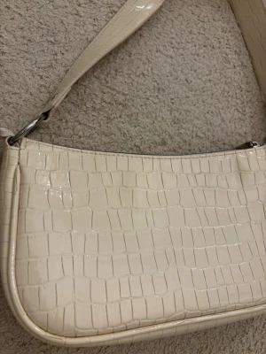 Krämvit axelväska i krokopräglat skinn - Säljer en stilren axelväska i krämvitt, med croco-mönstrad yta och bred axelrem. Väskan har en avlång form och stängs med dragkedja. Perfekt för dig som gillar trendiga accessoarer med lite extra attityd.