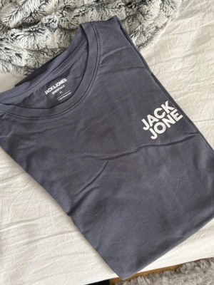 Mörkgrå t-shirt från Jack & Jones - Snygg mörkgrå t-shirt från Jack & Jones Essentials med vit logga på bröstet. Klassisk rund halsringning och normal passform. Tillverkad i mjuk bomull som känns skön mot huden. Perfekt basplagg till garderoben.