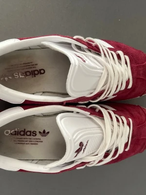Adidas Gazelle vinröda sneakers - Säljer ett par Adidas Gazelle sneakers i vinrött mocka med klassiska vita ränder och vit sula. Skorna har vita snören och detaljer i vitt på häl och plös. Perfekta för dig som gillar retrostil och vill sticka ut med färg. Ikonisk Adidas-logga på plösen. Skorna är knappt använda, det ända man kan se är lite smuts på sulan.