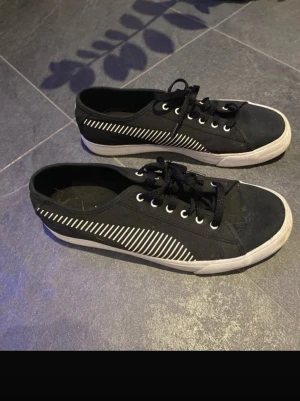Svarta sneakers med vita detaljer pumma - Snygga svarta sneakers från Pumma med vita ränder på sidorna och vita sulor. Skorna har klassisk låg design, svarta skosnören och metallöljetter. Tillverkade i canvasmaterial som ger en avslappnad och trendig look.