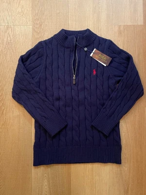 Marinblå kabelstickad tröja Polo Ralph Lauren - Snygg marinblå kabelstickad tröja från Polo Ralph Lauren med halv dragkedja och ribbad krage. Tröjan har det klassiska röda Polo-logot broderat på bröstet och är gjord i mjukt material. Perfekt för en stilren och avslappnad look.