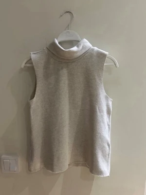 Beige polotopp från Zara - Säljer en stilren beige polotopp från Zara. Toppen är ärmlös och har en hög krage som ger en clean och modern look. Perfekt att matcha med jeans eller kjol för en trendig vibe.