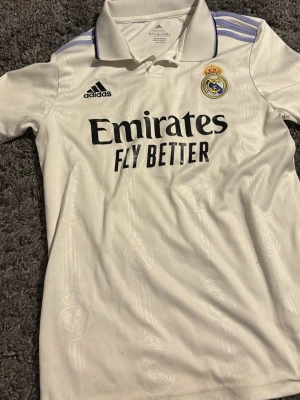 Real Madrid matchtröja 22/23 barn - Säljer en vit Real Madrid matchtröja från Adidas med klubbmärke och sponsortryck på bröstet. Tröjan har krage, svarta detaljer och lila ränder på axlarna. Pris kan diskuteras! Barnstorlek