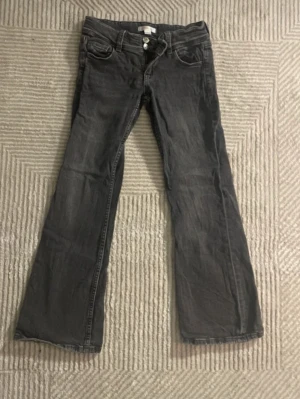 Grå bootcut jeans med dubbla knappar - Säljer ett par grå bootcut jeans med dubbla knappar i midjan och klassiska fem fickor. Jeansen har en lätt tvättad look och vida ben. Perfekta för dig som gillar retrostil och vill ha ett par bekväma jeans med snygg passform.