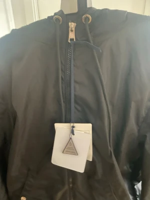 Svart vindjacka med huva - Snygg svart vindjacka med dragkedja framtill och justerbar huva med snörning. Jackan har fickor med tryckknappar och metalliska detaljer vid snörningen. Perfekt för blåsiga dagar och enkel att matcha med olika outfits.