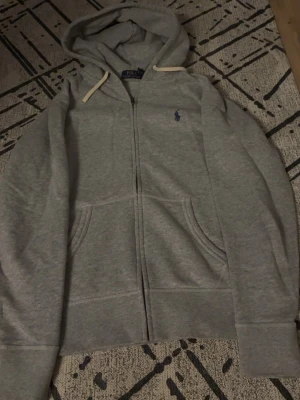 Grå hoodie från Polo Ralph Lauren - Snygg grå hoodie med dragkedja från Polo Ralph Lauren. Klassisk design med huva och vita snören, två fickor framtill och broderad logga på bröstet. Perfekt för en chill och avslappnad stil.