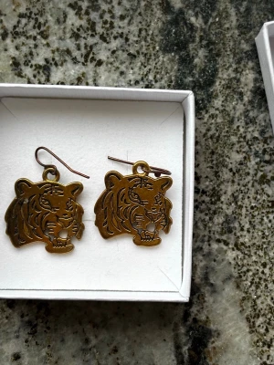 Guldiga örhängen med tigerhuvud - Coola örhängen i guldfärgad metall med detaljerat tigerhuvud som motiv. Hängande modell med krok, perfekt för dig som vill sticka ut med ett djurinspirerat smycke. Säljs i originalask.