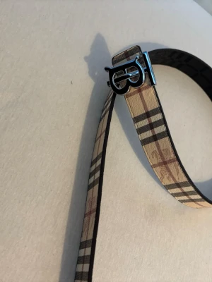 Rutmönstrat bälte från Burberry - Snyggt bälte från Burberry med klassiskt rutmönster i beige, svart och rött. Bältet har ett stilrent metallspänne med Burberrys logga och är tillverkat i skinn. Perfekt accessoar för att lyfta din outfit.