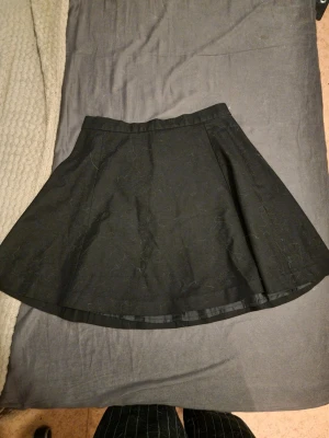 Svart kortkjol med lätt utsvängd form - Snygg svart kortkjol med lätt utsvängd siluett. Kjolen har en enkel design utan synliga detaljer och är fodrad på insidan för extra komfort. Perfekt att styla med en cool topp eller stickad tröja.