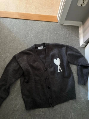 Mörkgrå cardigan från Ami Paris - Mysig mörkgrå cardigan från Ami Paris med v-ringning och knäppning framtill. På bröstet finns ett vitt hjärta och bokstaven A, typiskt för märket. Tröjan har ribbade muddar och är stickad i mjukt material, perfekt för lager på lager.